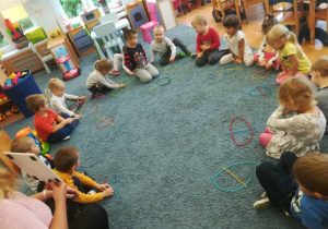Dzieci siedzą na dywanie w kole, układają z dwóch kolorowych sznurków wzór – jajko pośrodku którego z drugiego sznurka układają linię poziomą (koszyk)